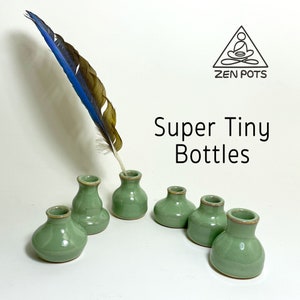 May include: Six small, sage green ceramic bottles with a light brown rim are arranged on a white surface. A blue and brown feather quill is inserted into one bottle. The text "Super Tiny Bottles" is displayed.