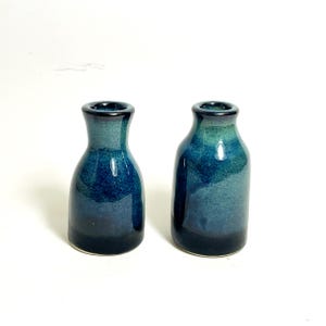 May include: Two small, ceramic bottles with a dark blue and teal glaze. The bottles have a rounded body and a narrow neck. The glaze has a glossy finish. The bottles are displayed against a white background.