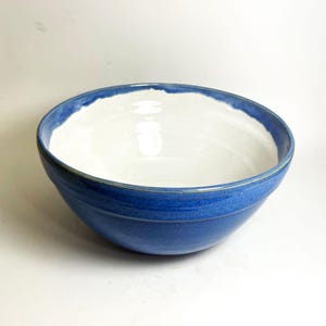 May include: A handmade ceramic bowl with a wide, round shape. The exterior is a vibrant blue, while the interior is white. The rim has a slightly uneven edge, adding to its unique, handcrafted appeal. This bowl is perfect for serving or display.