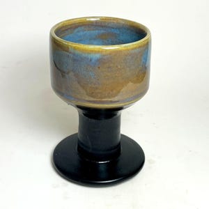 May include: A handmade ceramic goblet with a wide, cylindrical bowl and a black stem and base. The bowl features a blend of blue, brown, and gold hues, creating a unique, artistic appearance. The goblet is suitable for beverages.