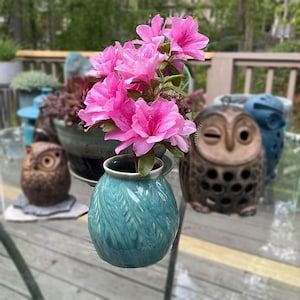 May include: A teal ceramic vase with a leaf pattern holds vibrant pink azalea flowers. The vase sits on a glass table, with decorative owl figurines and potted plants in the background. A charming floral arrangement.