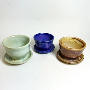 May include: Three handmade ceramic bowls with matching saucers. The bowls are in shades of green, blue, and brown. The bowls are round with a slightly flared rim. The saucers are flat and round. The bowls and saucers are glazed with a glossy finish.