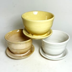 May include: Three ceramic bowls with matching saucers, arranged in a cluster. One bowl is bright yellow, one is a soft beige with subtle horizontal ridges, and one is a clean white. Each bowl sits securely on its own circular saucer.
