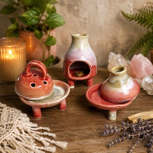 May include: Three ceramic incense burners with a glossy finish in shades of pink, cream, and red. Each piece features a rounded base with three small legs, a removable lid with ventilation holes, and a smooth, glazed surface texture.