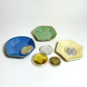 May include: A collection of small, hexagonal ceramic dishes in blue, green, and yellow, each holding a coin in a protective case. The coins are gold and silver, with detailed designs.