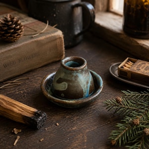 May include: A handcrafted ceramic incense holder with a teal and brown glaze. The holder sits on a matching saucer, perfect for catching ash. The rustic design is complemented by a vintage book and a box of matches, creating a cozy, natural aesthetic.