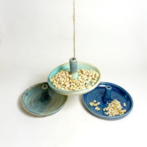 May include: Three handmade ceramic bird feeders in shades of blue and green. Each feeder has a circular dish with a central post and is filled with seeds. One feeder is suspended by a twine string.