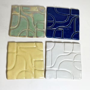 May include: Four square ceramic tiles in shades of green, blue, yellow, and white. Each tile features a raised, abstract design of curved and angular lines. The tiles are arranged on a white surface, showcasing their varied colors and textures.