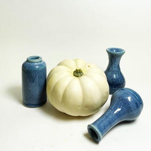 May include: A still life arrangement featuring a white pumpkin and three small blue ceramic vases. The vases have a speckled glaze. The pumpkin is round with a green stem.