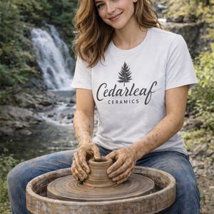May include: A person wearing a white t-shirt with the text "Cedarleaf Ceramics" and a tree graphic, working on a pottery wheel. The person is wearing blue jeans and is in an outdoor setting with a waterfall in the background.