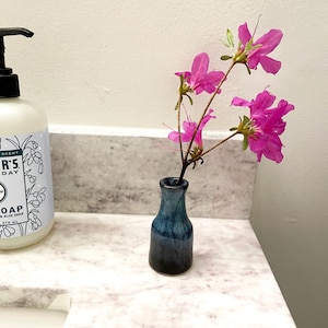 May include: A small, blue and black ceramic vase holds a sprig of bright pink flowers. The vase has a glossy finish and a narrow neck. The flowers have multiple petals and are in full bloom. The vase is on a white countertop.