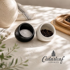 May include: A black ceramic salt cellar filled with white salt and a white ceramic pepper cellar filled with black peppercorns. The salt and pepper cellars are egg-shaped and sit on a light-colored surface. The Cedarleaf Ceramics logo is visible.