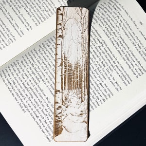 Può includere: Un segnalibro in legno con un design inciso a laser di un sentiero boschivo innevato. Il sentiero si snoda attraverso un boschetto di betulle su uno sfondo marrone chiaro.