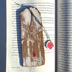 May include: A wooden bookmark with a detailed illustration of a cathedral, featuring a red accent. A small, round charm with a pink rose hangs from the top. The bookmark is placed on an open book.