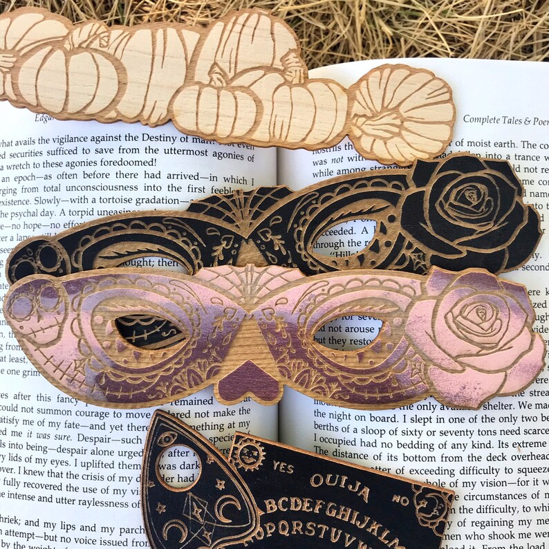 May include: A set of four wooden bookmarks featuring a black rose, a pink and purple butterfly mask, and a pumpkin design. The bookmarks are layered on top of a book with the text "Complete Tales & Poems" visible. The bookmarks are perfect for adding a touch of whimsy to your reading experience.