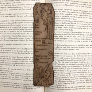 May include: A wooden bookmark with a fairy design. The fairy is holding a book and has a quote about weeping. The bookmark is brown and has a natural wood finish.