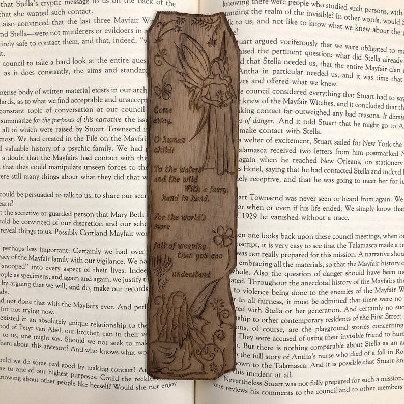 May include: A wooden bookmark with a fairy design. The fairy is holding a book and has a quote about weeping. The bookmark is brown and has a natural wood finish.