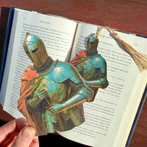 May include: A unique bookmark featuring a knight in shining armor holding a red rose, set against the backdrop of an open book. The bookmark has a gold tassel. The book pages are visible.