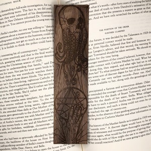 May include: A wooden bookmark with a laser-engraved design of a skull with tentacles and a pentagram. The bookmark is brown and has a natural wood grain. The text "The Position of the Order Further Clarified" is visible on the page behind the bookmark.