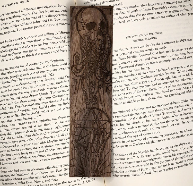 May include: A wooden bookmark with a laser-engraved design of a skull with tentacles and a pentagram. The bookmark is brown and has a natural wood grain. The text "The Position of the Order Further Clarified" is visible on the page behind the bookmark.