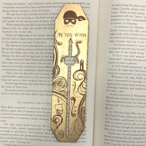 May include: A wooden bookmark with a gold-colored design. The bookmark features a silhouette of a person wearing a mask and holding a sword. The text "As You Wish" is written on the bookmark.