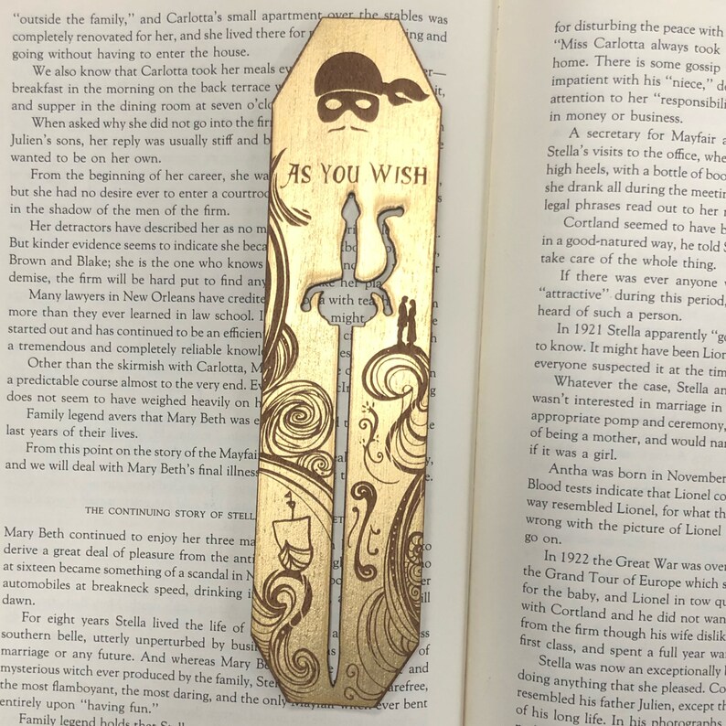 May include: A wooden bookmark with a gold-colored design. The bookmark features a silhouette of a person wearing a mask and holding a sword. The text "As You Wish" is written on the bookmark.