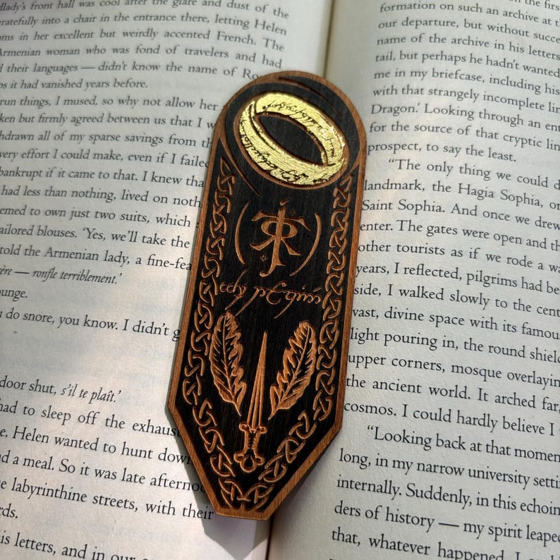 May include: A wooden bookmark with a black and gold design. The bookmark features a ring with an inscription, a sword, and leaves. The inscription reads "The One Ring" and "One Ring to rule them all, One Ring to find them, One Ring to bring them all and in the darkness bind them."