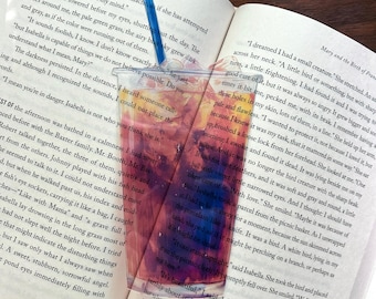 Puede incluir: Primer plano de un libro abierto con un marcapáginas colorido. El marcapáginas es una taza de plástico transparente llena de un líquido rojo. El libro está abierto en una página con texto sobre un sueño. El texto dice: "Soñé que tenía una pequeña criatura."