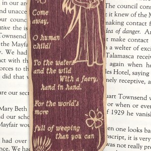May include: A wooden bookmark with a whimsical illustration of a fairy with wings and a flower. The bookmark features the text "Come away, O human child! To the waters and the wild With a faery, hand in hand. For the world's more full of weeping than you can understand."