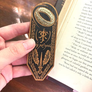 May include: A wooden bookmark with a black and gold design. The bookmark features a ring, a sword, and the text "(來) est".