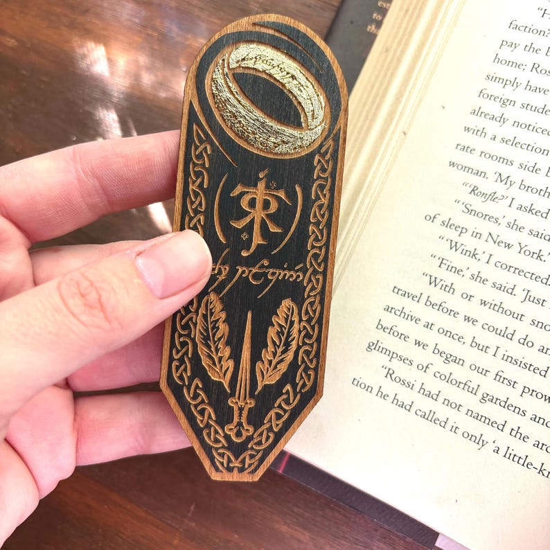 May include: A wooden bookmark with a black and gold design. The bookmark features a ring, a sword, and the text "(來) est".