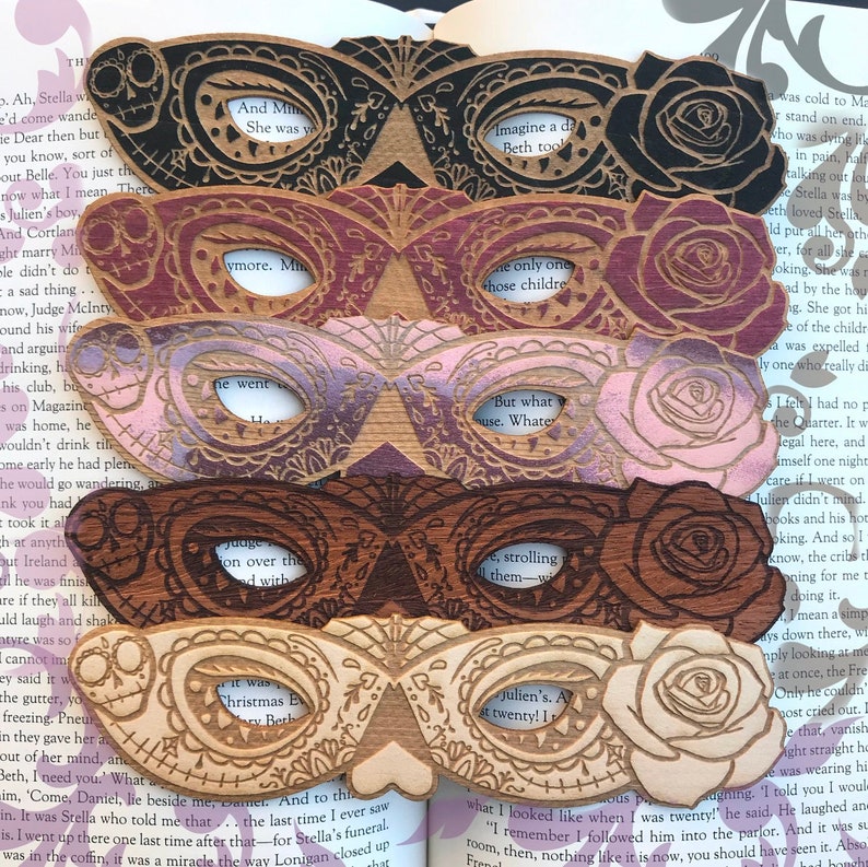 May include: Five wooden laser-cut masks with intricate designs, each featuring a rose and a different color scheme: black, burgundy, purple, brown, and natural wood. The masks have text cut out of them, including "And Mimi", "Imagine a day Beth took", "the only one those children", "But what use. What", and "Julien's. At twenty! I".