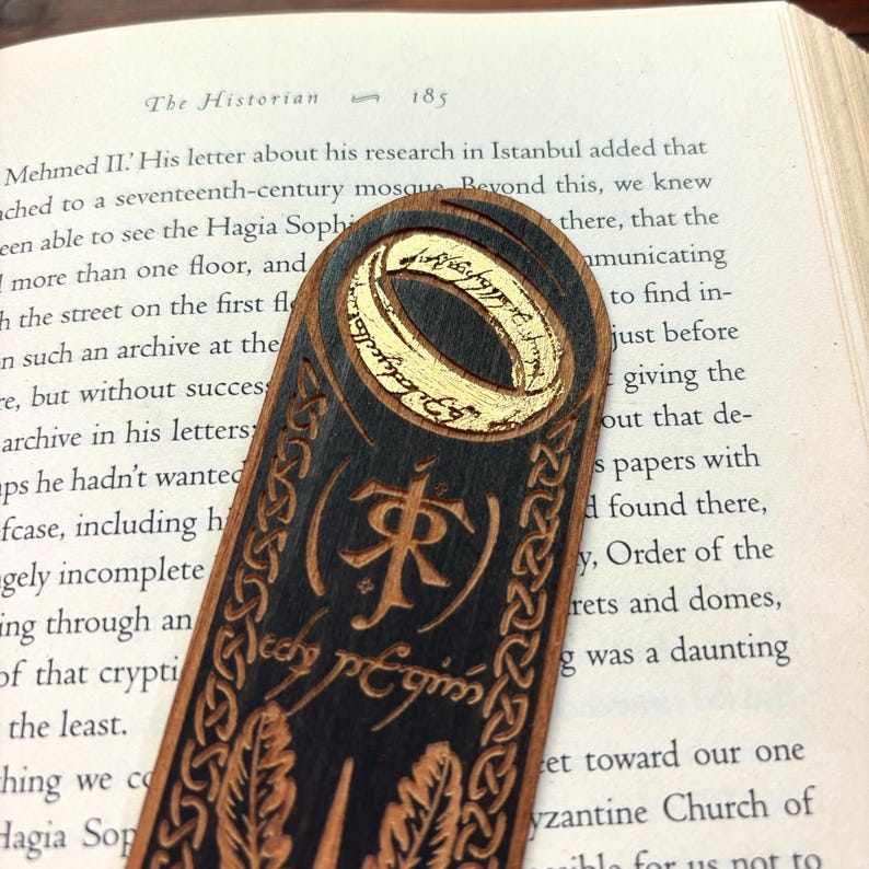 May include: A wooden bookmark with a gold ring design and the inscription "One Ring to rule them all" in Elvish script. The bookmark is black with a dark wood grain and features a decorative border.
