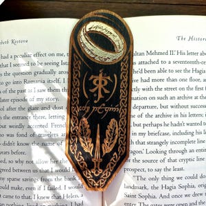 May include: Wooden bookmark with a black background and a silver ring with the inscription "One Ring to rule them all" in white. The bookmark also features a black and gold inscription in a stylized font, a sword, and two leaves.