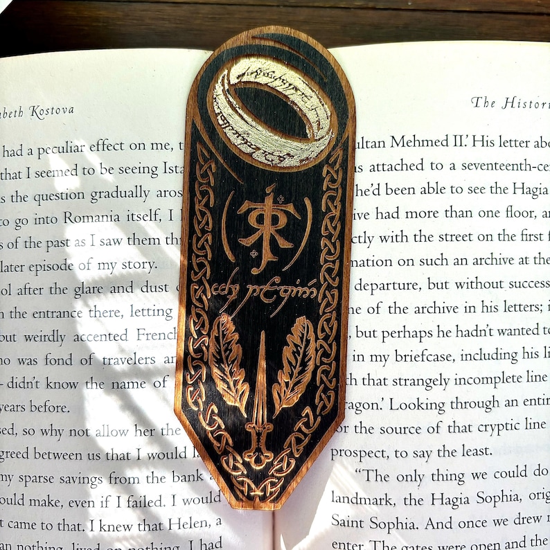 May include: Wooden bookmark with a black background and a silver ring with the inscription "One Ring to rule them all" in white. The bookmark also features a black and gold inscription in a stylized font, a sword, and two leaves.