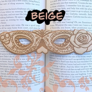 May include: A wooden mask with intricate carvings and a beige background. The mask is shaped like a masquerade mask with a heart-shaped opening in the center. The mask is resting on an open book with text visible.