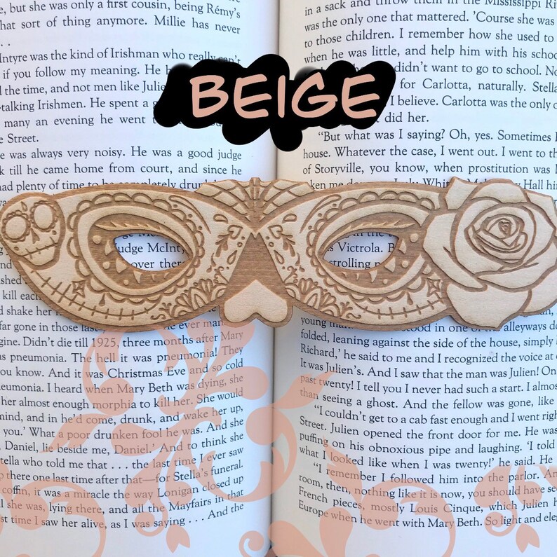May include: A wooden mask with intricate carvings and a beige background. The mask is shaped like a masquerade mask with a heart-shaped opening in the center. The mask is resting on an open book with text visible.