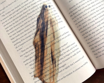 May include: A close-up of a book page with a brown and white bookmark shaped like a ghost. The bookmark is made of wood and has a textured surface. The text on the page reads "He won't drown you, he won't drag you to the bottom of the sea, he won't rip you open with his yellow claws and yet he's one of the most dangerous creatures out there..."