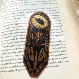 May include: A wooden bookmark with a black background and gold accents. The bookmark features a ring with the inscription "One Ring to rule them all, One Ring to find them, One Ring to bring them all and in the darkness bind them." The bookmark also features a sword and two feathers, and the inscription "The One Ring" in Elvish.