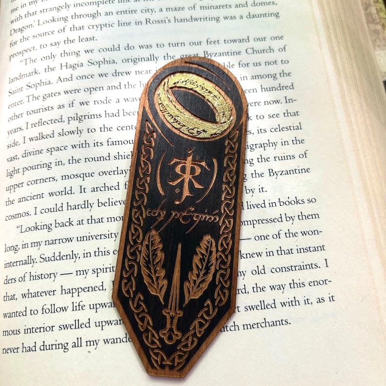 May include: A wooden bookmark with a black background and gold accents. The bookmark features a ring with the inscription "One Ring to rule them all, One Ring to find them, One Ring to bring them all and in the darkness bind them." The bookmark also features a sword and two feathers, and the inscription "The One Ring" in Elvish.