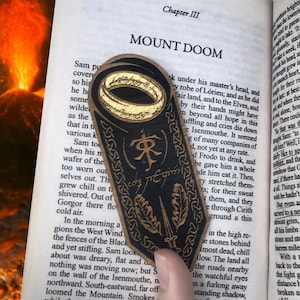 May include: A black leather bookmark with a gold ring, a sword, and leaves. The bookmark has the inscription "One Ring to rule them all" and the text "Mount Doom" is visible in the background.