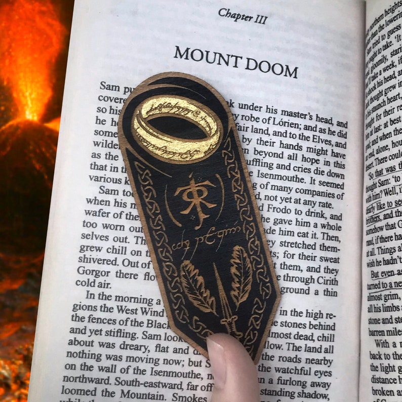 May include: A black leather bookmark with a gold ring, a sword, and leaves. The bookmark has the inscription "One Ring to rule them all" and the text "Mount Doom" is visible in the background.