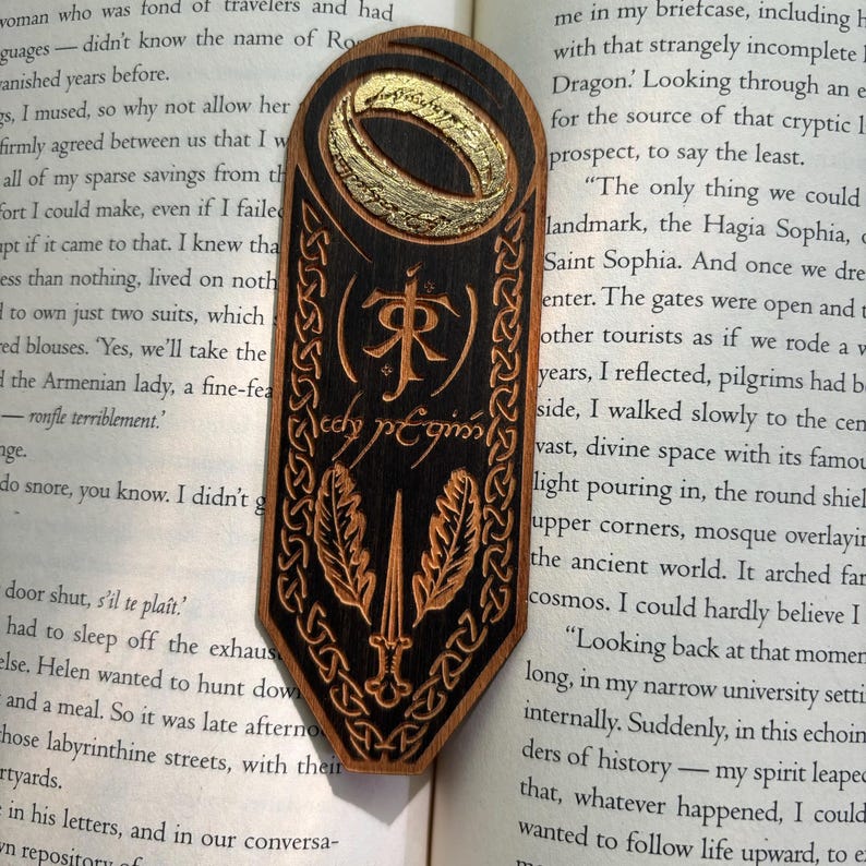 May include: A wooden bookmark with a gold-painted ring, a black and brown design, and the inscription "The One Ring" in Elvish script. The bookmark is shaped like a shield with a sword and leaves.