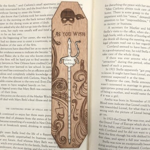 May include: A wooden bookmark with a laser-cut design of a sword and a couple standing on a balcony. The bookmark is brown and has a light wood grain. The text "As You Wish" is printed on the bookmark.