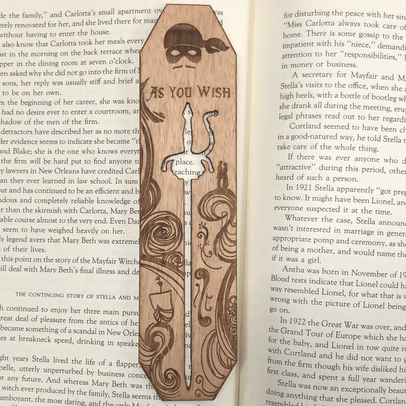 May include: A wooden bookmark with a laser-cut design of a sword and a couple standing on a balcony. The bookmark is brown and has a light wood grain. The text "As You Wish" is printed on the bookmark.