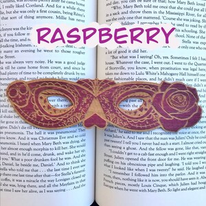 May include: A wooden bookmark shaped like a masquerade mask with a raspberry design. The bookmark has a cutout in the center with the word "Raspberry" written in purple letters.