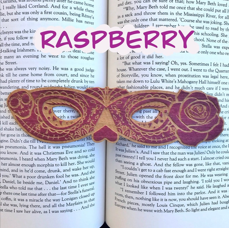 May include: A wooden bookmark shaped like a masquerade mask with a raspberry design. The bookmark has a cutout in the center with the word "Raspberry" written in purple letters.