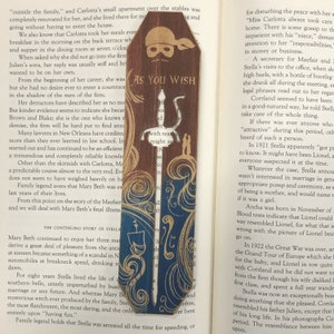 May include: A wooden bookmark with a blue and gold design. The bookmark features a pirate skull and crossbones with the words "As You Wish" written on it. The bookmark is decorated with a sword and a swirling pattern.