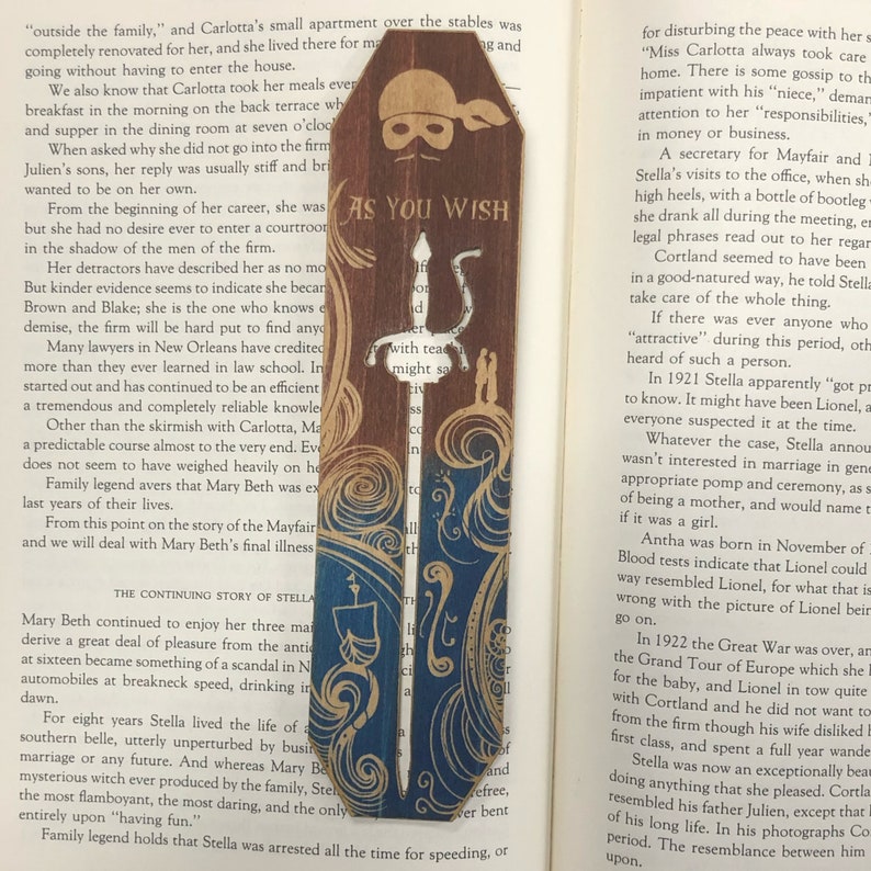 May include: A wooden bookmark with a blue and gold design. The bookmark features a pirate skull and crossbones with the words "As You Wish" written on it. The bookmark is decorated with a sword and a swirling pattern.