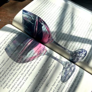 Op de afbeelding: Een close-up van een boek met een glas wijn erop. Het glas wordt gedeeltelijk bedekt door het boek, maar de wijn erin is diep rood. Het boek is open op een pagina met tekst, en het licht van een raam schijnt door de pagina's.